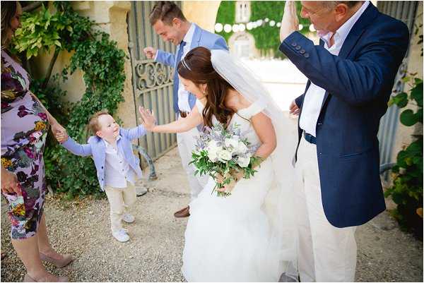 A candid moment captured outdoors at what appears to be a French chateau or manor, where a bride in a white ballgown with a tulle veil bends down to high-five a young boy dressed in a light blue vest and khaki trousers. The bride holds a bouquet of white roses, anemones, and greenery with purple thistle accents. Several guests are visible around her, including a man in a navy blue suit and a woman in a floral dress, with a second man in a light blue suit visible in the background near an ornate iron gate. The setting is a gravel courtyard with ivy-covered stone walls and string lights overhead. Wide candid shot with a warm, natural daylight feel.