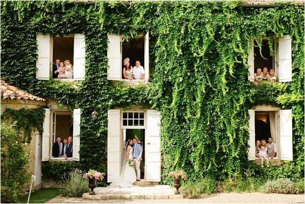 A wide shot of a two-storey French country house with a facade densely covered in climbing green ivy, featuring white-shuttered windows and a central doorway. The bride, in a white gown, and groom, in a light blue suit, stand together at the front entrance steps, with potted pink floral arrangements flanking the doorway. Wedding guests — approximately 15 to 20 people — are distributed across multiple windows on both floors, leaning out and looking toward the camera in a playful, staged group portrait composition. Potential venue feature image.