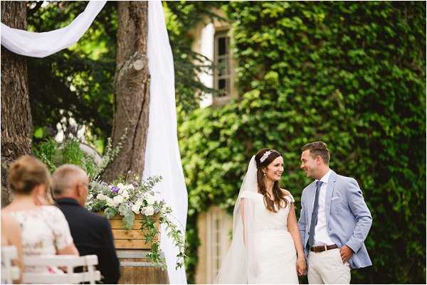 An outdoor wedding ceremony taking place in a chateau courtyard with ivy-covered stone walls and large trees. The bride, wearing a white gown with a cathedral-length veil and a floral headpiece, and the groom, dressed in a light blue blazer with a grey tie and beige trousers, stand together smiling at each other at the altar. A white fabric drape is wrapped around a tree trunk behind them, and a wooden barrel holds a floral arrangement featuring white blooms, purple flowers, and eucalyptus foliage. Two seated guests are visible in the foreground on white chairs, viewed from behind. The composition is a medium-wide portrait shot capturing both the couple and elements of the ceremony setup.