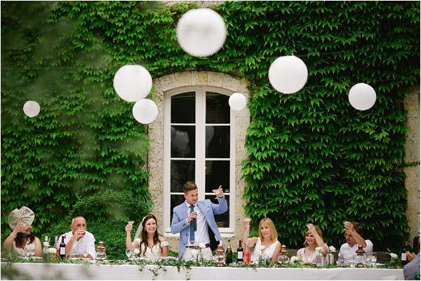 A groom in a light blue suit delivers a speech at an outdoor reception head table, holding a microphone in one hand and gesturing with the other, while approximately eight guests seated around him listen and raise glasses in a toast. The table is dressed with a white linen, scattered greenery, and various bottles and glassware. The setting is in front of a stone building facade heavily covered in climbing ivy, with a single arched window visible behind the group. Six large white round paper lanterns or balloon spheres are suspended in the air above the table, adding a festive decorative element to the scene. Wide shot composition capturing the full table and architectural backdrop.