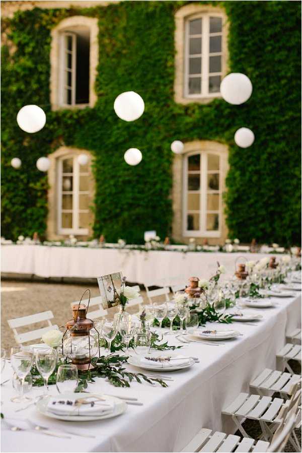 An outdoor wedding reception table setup photographed in a wide shot against the ivy-covered stone facade of what appears to be a French chateau. A long rectangular banquet table is dressed in a white linen tablecloth and lined with white folding chairs, set for approximately 15-20 guests. The centerpiece runner consists of lush greenery — trailing foliage and leafy branches — interspersed with copper lanterns, white floral accents, small bud vases, and framed place cards or menus. Place settings include stacked white plates, multiple wine glasses, and laid cutlery. Overhead, a cluster of white globe pendant lights hang at varying heights in front of the ivy wall, creating a warm, lit atmosphere as evening approaches. The decor palette is white, green, and copper, with a classic-meets-rustic styling. Potential venue feature image.