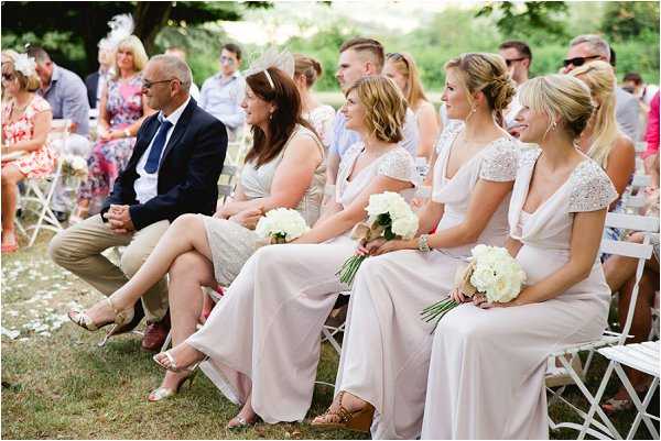 An outdoor wedding ceremony is underway, with guests and bridesmaids seated on white folding chairs on a lawn. Three bridesmaids are prominently visible in the foreground, wearing floor-length blush pink dresses — two with lace cap sleeves — each holding bouquets of white peonies or ranunculus. The wider guest seating includes casually dressed attendees in summer clothing, with one woman wearing a fascinator. The shot is a medium wide-angle taken from a slight angle, capturing the relaxed, garden-party atmosphere of the ceremony in what appears to be a French outdoor setting with trees in the background.