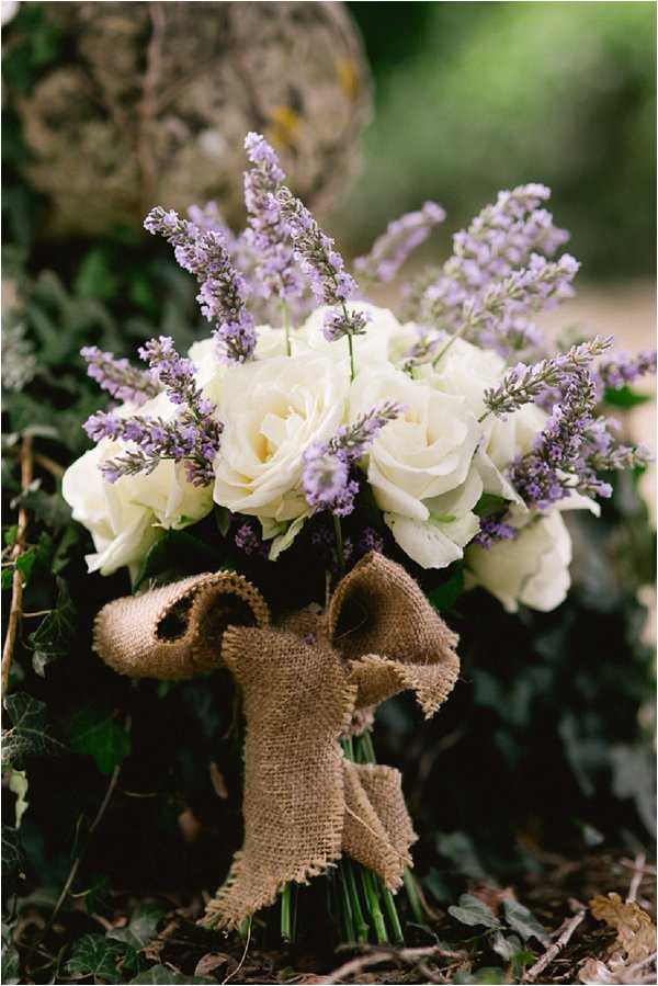 Close-up detail shot of a bridal bouquet resting on the ground among ivy leaves outdoors. The bouquet is composed of cream/ivory roses and stems of purple lavender, tied with a large burlap bow in a rustic style. The stems are bound together and the burlap ribbon features frayed edges, reinforcing the rustic, countryside aesthetic. The background is softly blurred with greenery, keeping the focus entirely on the bouquet.