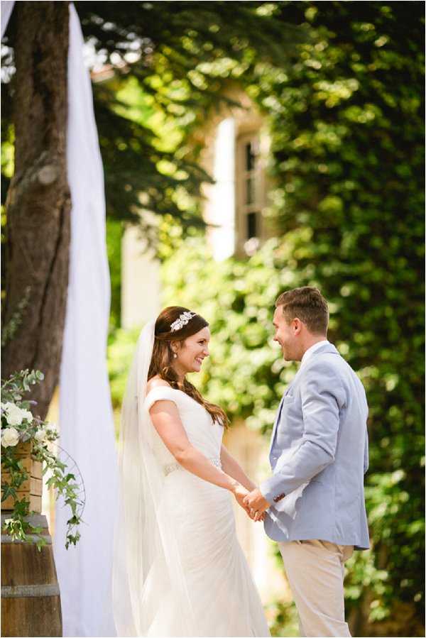 A bride and groom hold hands and face each other during an outdoor wedding ceremony, appearing to exchange vows or share a moment of laughter. The bride wears a fitted ivory gown with cap sleeves and a long veil secured with a crystal headband, while the groom is dressed in a light blue suit jacket with beige trousers. The ceremony is set in a garden or estate courtyard, with a white fabric drape and a wooden barrel topped with white floral arrangements including white blooms and trailing greenery visible to the left. A ivy-covered stone building is softly blurred in the background. The composition is a medium portrait shot with shallow depth of field.