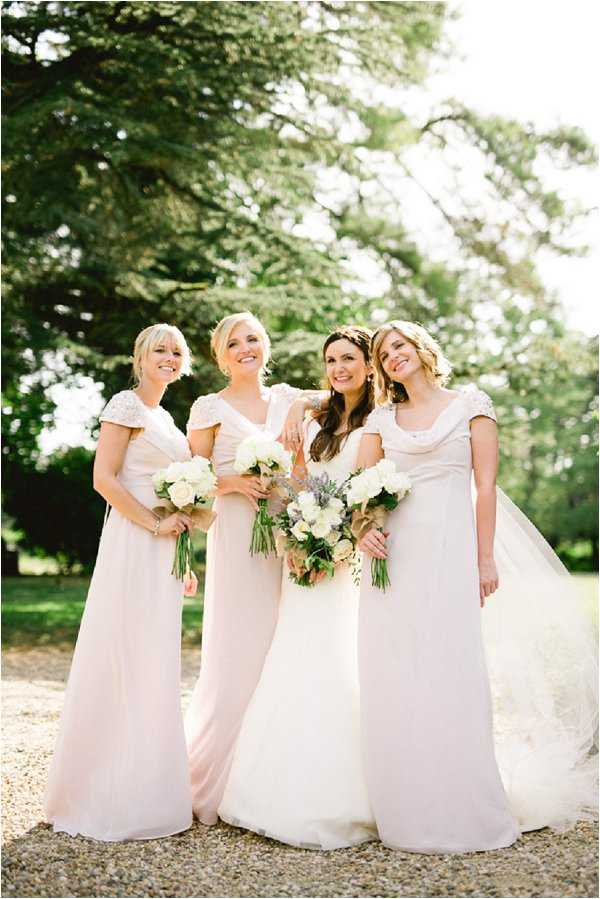 A bridal party portrait taken outdoors on a gravel path, featuring a bride and three bridesmaids posing together in a medium-wide shot. The bride wears a white A-line gown with a long veil and a floral hair accessory, and carries a loose bouquet of white and cream blooms with lavender and greenery. The three bridesmaids wear floor-length blush pink dresses with cap sleeves featuring subtle embellishment or ruching, each holding small rounded bouquets of white flowers including what appear to be white roses and hydrangeas with green stems. The overall styling is soft and romantic with a classic, understated palette of white and blush. Natural backlighting creates a bright, airy quality to the image.