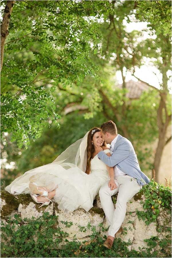 A couple portrait taken outdoors, with the bride and groom sitting together on a low stone wall covered in trailing ivy. The bride wears a strapless ivory ballgown with a beaded waist detail, a long cathedral-length veil, a small floral hair piece, and white strappy heeled shoes. The groom is dressed in a light blue blazer, white trousers, and tan loafers. He leans in to kiss her cheek while she laughs and looks toward him. The setting has a rustic Provençal feel with dappled warm light filtering through the tree canopy above. The shot is a medium-wide portrait with soft natural bokeh in the background where a tiled rooftop is faintly visible.