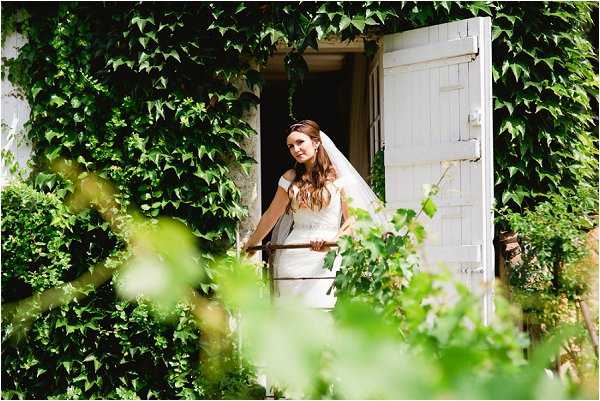 A bridal portrait showing the bride standing in a white wooden doorway of a ivy-covered stone building, looking outward. She is wearing a fitted off-the-shoulder white gown with a beaded waist detail and a long veil, with her dark auburn hair worn down. The shot is framed through foreground foliage, creating a layered, partially obscured composition that draws the eye to the bride in the doorway. The overall styling has a classic, natural feel with the white shuttered door and dense green ivy covering the building facade.