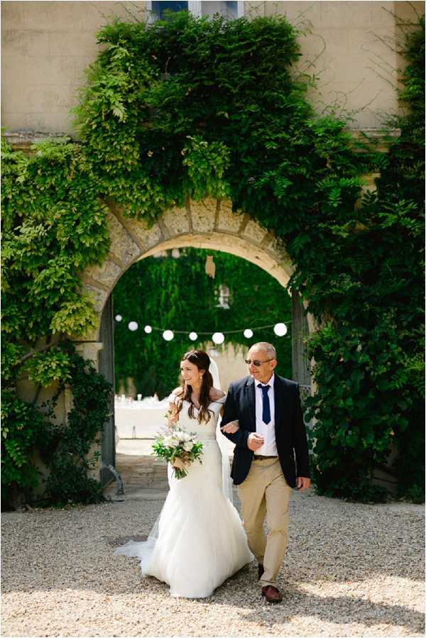 A bride is being escorted through a large stone archway covered in dense green climbing foliage, likely at a French chateau or manor house. She is wearing a white fit-and-flare gown with a long tulle skirt and veil, and carries a loose, garden-style bouquet with soft white and blush blooms and greenery. The man accompanying her wears a navy blazer, navy tie, khaki trousers, and sunglasses. A strand of white paper lanterns is strung across the archway in the background, and the ground is covered in pale gravel. The shot is a medium full-length portrait taken outdoors in bright daylight.