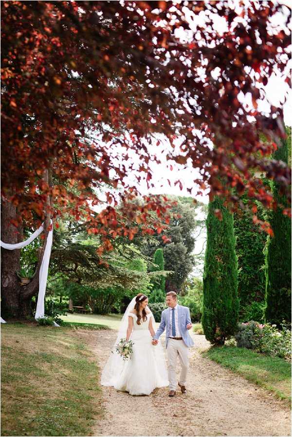 A bride and groom walk hand-in-hand along a gravel garden path during couple portraits taken outdoors. The bride wears a white ballgown with a cap-sleeved lace bodice and a cathedral-length veil, carrying a loose bouquet of white and greenery blooms, while the groom wears a light blue blazer, khaki trousers, a blue tie, and brown leather shoes. The path is framed by deep burgundy-leafed trees overhead and tall cypress trees lining the sides, with white fabric draping visible on a tree trunk to the left, suggesting a nearby ceremony setup. The composition is a medium full-length portrait shot from a low angle, capturing the relaxed, candid mood of the pair laughing as they walk toward the camera.