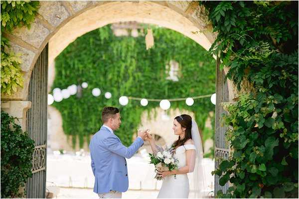 A couple portrait taken outdoors at a French chateau or manor, with the bride and groom framed by a stone archway covered in climbing green ivy. The groom wears a light blue suit and is placing or adjusting something on the bride's hand or wrist, and both are smiling at each other. The bride wears an off-the-shoulder white fitted gown with a cathedral veil and a pearl or crystal headband, and holds a bouquet of white blooms with lavender and eucalyptus greenery. White paper lantern string lights are strung across the courtyard visible through the archway in the background, suggesting a relaxed, classic outdoor reception setup. The shot is a medium portrait with soft natural light and slightly blurred background depth.