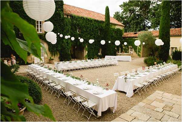 An outdoor wedding reception setup in the courtyard of a French stone manor house covered in ivy, with a terracotta-tiled roof. Several long rectangular tables are arranged in a U-shape layout, dressed with white linen tablecloths, white bistro-style folding chairs, and centerpieces featuring greenery and small pink floral accents with glass bottles. White paper lanterns are strung across the courtyard on lines above the tables, adding decorative overhead interest. The wide-angle shot captures the full table arrangement and venue facade, with a gravel courtyard surface and cypress trees visible in the background. Potential venue feature image.