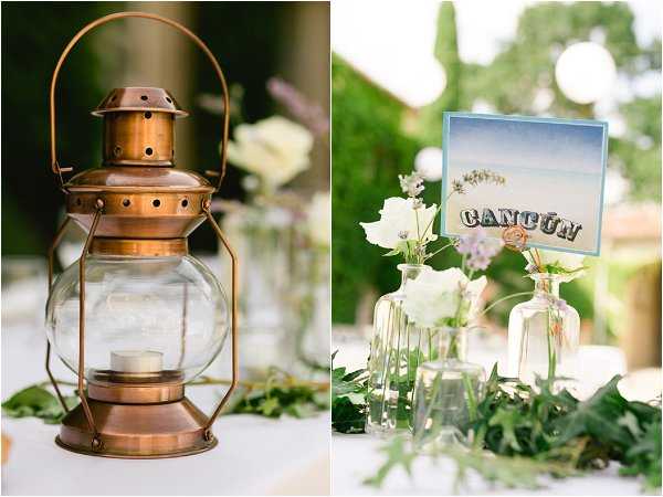 Close-up detail shots of outdoor wedding reception table decor presented as a side-by-side diptych. On the left, a copper-toned vintage lantern with a glass globe holds a small tealight candle, set against a backdrop of trailing greenery on a white tablecloth. On the right, a travel-themed table number card reading 'CANCÚN' with a postcard-style design is displayed among small glass bud vases and test tube vases holding single white roses and delicate wildflowers, with scattered greenery. The overall decor theme is vintage travel, featuring a warm copper and white color palette with natural greenery accents.
