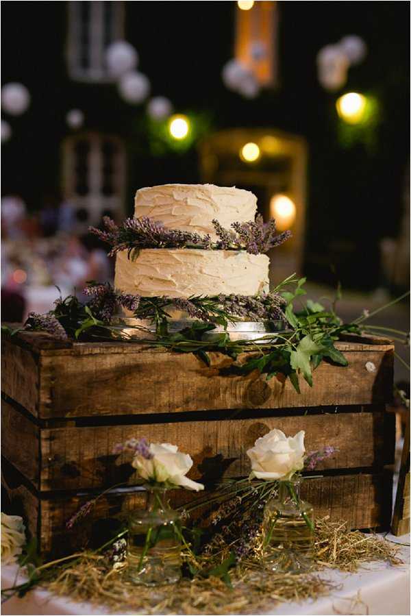 Close-up detail shot of a two-tier wedding cake with textured ivory buttercream frosting, decorated between layers with fresh lavender sprigs and greenery. The cake is displayed on a rustic wooden crate, with additional lavender bunches, white roses in small glass bud vases, and dried straw arranged around the base, creating a rustic, Provençal-style display. The setting appears to be an outdoor evening reception, with warm string lights and what appear to be white paper lantern decorations visible in the blurred background, along with the lit facade of what appears to be a stone building. The overall decor palette combines ivory, purple lavender, and natural wood tones in a rustic country style.