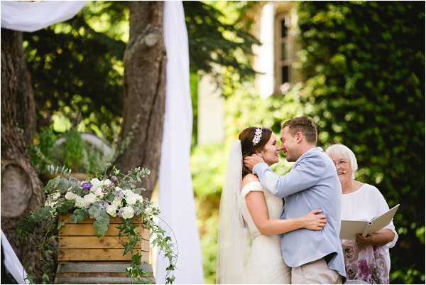 The bride and groom share their first kiss during an outdoor ceremony, with the officiant standing beside them holding a clipboard. The ceremony takes place in a garden setting with ivy-covered stone building architecture visible in the background. The bride wears a white off-shoulder gown with a cathedral veil and a floral hair piece, while the groom is dressed in a light blue suit. A wooden crate serves as a ceremony decoration, topped with an arrangement of white roses, white anemones, purple flowers, and trailing eucalyptus foliage, with white fabric draping framing the scene. The shot is a medium portrait-style composition capturing the couple's embrace from a slight side angle.