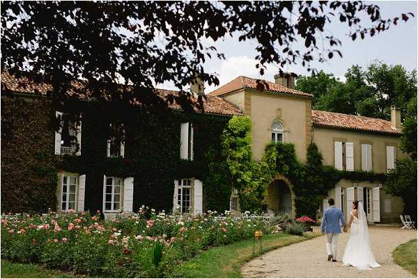 A couple walks hand-in-hand along a gravel path toward a large ivy-covered French country manor with terracotta roof tiles, white shutters, and a stone archway entrance. The bride wears a long white gown with a veil, and the groom is dressed in a light blue suit. A rose garden with pink and coral blooms lines the path in the foreground. The wide shot captures the full facade of the property surrounded by mature trees and climbing greenery. Potential venue feature image.