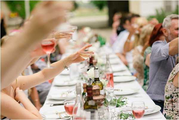 Guests seated at a long rectangular outdoor dining table raise their glasses in a toast during a wedding reception. The table is set with white plates, wine glasses containing rosé, green foliage garland running down the center, and wine bottles with small floral arrangements of white blooms. Approximately 15–20 guests are visible along both sides of the table, dressed in summer attire including floral and patterned outfits. The setting appears to be a shaded outdoor terrace or courtyard. The composition is a medium wide shot taken from a low angle at table level, with the foreground slightly blurred from shallow depth of field, giving focus to the raised glasses and table centerpieces.