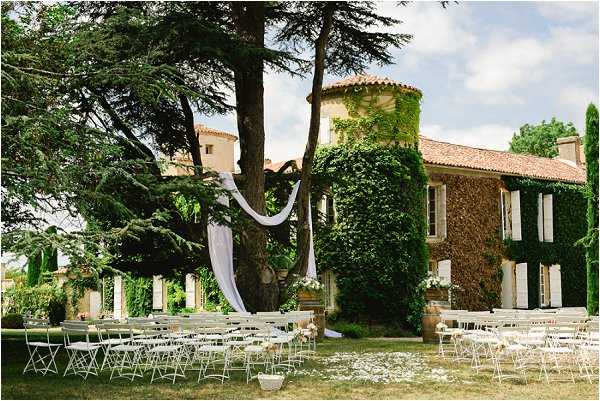 An outdoor wedding ceremony setup on the grounds of a French chateau or mas, with the ivy-covered stone building and a round tower visible in the background. White folding chairs are arranged in rows on either side of a central aisle, with small white floral arrangements placed along the aisle on the ground. A ceremony backdrop is created using white draped fabric tied to a large mature tree, with a wooden or rustic floral installation at its base featuring white blooms. The overall decor palette is white with a classic, understated style. Wide shot capturing the full ceremony space and venue facade. Potential venue feature image.