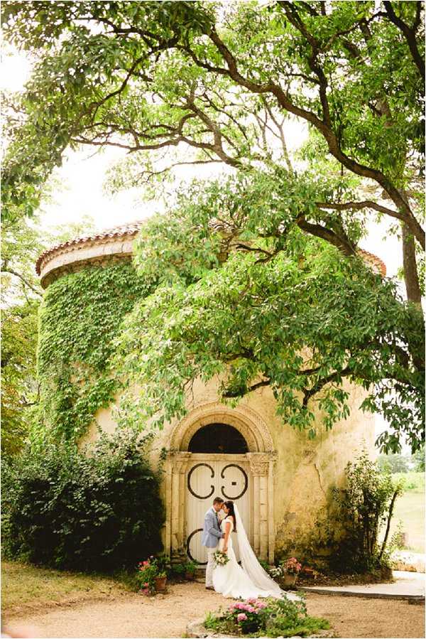 A couple shares a close moment standing in front of the ornate arched doorway of a small ivy-covered Romanesque chapel or chapel-style building, shot outdoors in natural daylight. The bride wears a white gown with a long cathedral veil and holds a bouquet with pink and white blooms, while the groom is dressed in a light grey suit. The building's stone facade is heavily covered in climbing green ivy, with a decorative carved stone surround framing the arched wooden door. The composition is a wide portrait shot framed by large tree branches overhead, with potted pink flowering plants visible at ground level near the entrance. Potential venue feature image.