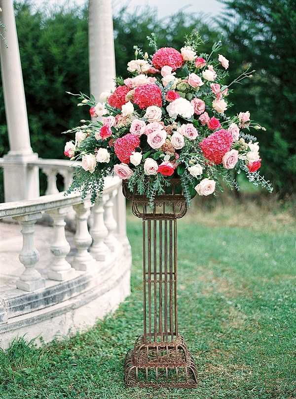 A close-up detail shot of a large floral arrangement displayed on a tall, ornate wrought-iron pedestal stand with a rust-brown patina. The arrangement features hot pink hydrangeas, blush and light pink garden roses, small coral spray roses, and trailing eucalyptus and mixed greenery. The overall palette is warm pink and coral with soft green accents. The pedestal is positioned on a lawn beside a curved white stone balustrade with classical columns, suggesting a formal chateau or manor house terrace setting. The floral style is lush and full, consistent with a classic or romantic wedding aesthetic.