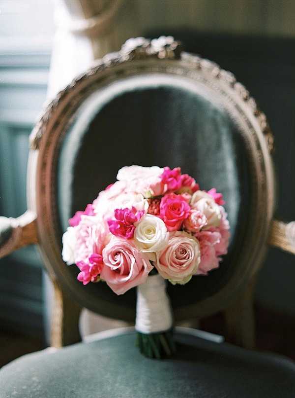 A close-up detail shot of a bridal bouquet resting on the seat of an ornate French Louis XVI-style armchair with a gold-trimmed oval back and dark teal upholstery. The bouquet is composed of hot pink roses, blush pink roses, cream roses, pink peonies, and magenta sweet peas or ranunculus, with stems wrapped in white ribbon. The color palette is bold, mixing deep fuchsia and soft blush tones. The chair is slightly out of focus in the background, drawing attention to the florals in the foreground.