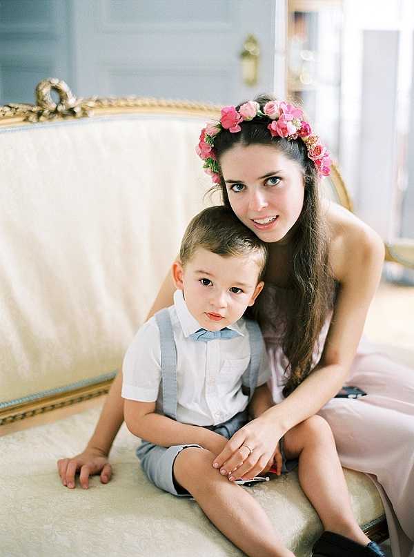 A portrait of a young woman and a small boy, approximately 3–4 years old, seated together on a gilded French-style cream upholstered settee in an indoor interior room. The woman wears a blush pink dress and a floral crown made of bright pink roses and greenery in her long dark hair, and has a ring on her finger. The boy is dressed in light grey shorts, grey suspenders, and a white short-sleeve shirt. The background features pale blue-grey panelled walls with gold detailing, suggesting a chateau or formal interior. The image is a medium portrait shot with soft, film-like exposure and warm tones.