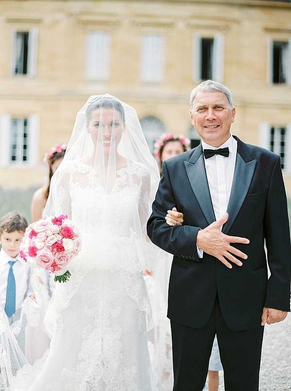 A bride is being escorted, likely by her father, in an outdoor processional in front of a large French chateau with pale stone façade visible in the background. The bride wears a long-sleeved lace wedding gown with a cathedral-length veil worn over her face, and carries a round bouquet of hot pink, blush, and ivory roses. Her escort wears a black tuxedo with a bow tie and appears to be smiling broadly toward the camera. In the background, bridesmaids wearing pink floral crowns are visible, along with a young boy in a white outfit with a blue tie. The composition is a mid-shot portrait framing the bride and her escort from approximately the knees up, with a classic, formal wedding style.