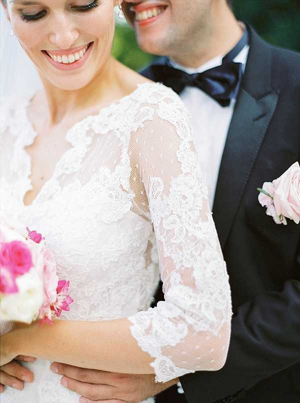 A close-up portrait of a bride and groom sharing a joyful moment together. The bride wears a white lace gown with three-quarter-length sheer lace sleeves featuring intricate floral lace detailing, and she holds a bouquet of hot pink and light pink roses. The groom is dressed in a dark navy suit with a matching navy bow tie and a light pink rose boutonniere on his lapel. The composition is a tight crop focusing on the couple from approximately the shoulders down to the waist, with both subjects smiling warmly.