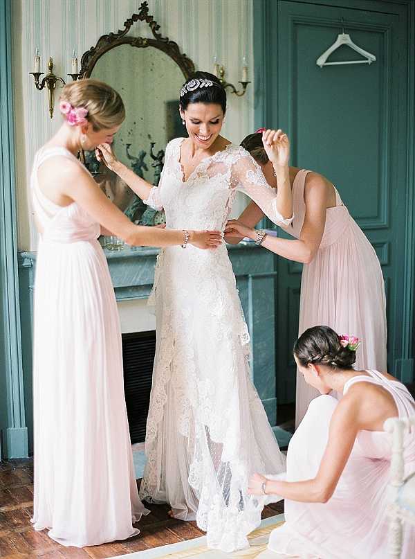 A getting-ready scene captured indoors, showing a bride being assisted by three bridesmaids as they fasten and adjust her gown. The bride wears a long-sleeved lace wedding dress with a v-neckline, tiered lace skirt, and a jeweled floral tiara, and is smiling broadly. The three bridesmaids wear floor-length blush pink dresses and have pink floral hair accessories — one stands on each side of the bride while a third kneels to adjust the hem. The room features teal-painted walls, an ornate gold-framed oval mirror above a marble fireplace mantel, and wall-mounted candle sconces, suggesting a French chateau interior. The shot is a medium full-length portrait with warm, soft natural lighting.