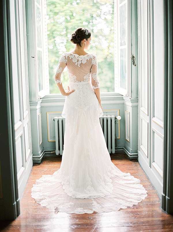 A bridal portrait taken indoors, showing the bride from behind as she stands facing tall French windows in what appears to be a chateau or historic venue interior. She wears an ivory lace wedding gown with three-quarter length illusion lace sleeves, a sheer lace bodice with appliqué floral detailing, and a scallop-edged cathedral train spread across warm hardwood floors. Her dark hair is styled in an updo with a small hair accessory. The room features pale sage-green painted woodwork, white double doors framing the shot, and a white cast-iron radiator visible to the right. The composition is a full-length portrait framed symmetrically by the doorway architecture, with soft natural window light illuminating the scene.