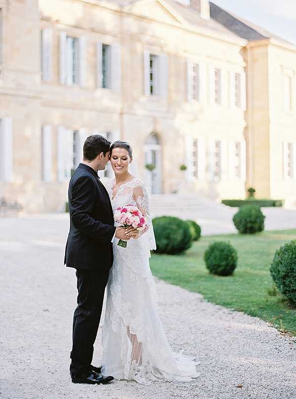 A couple portrait taken outdoors on the gravel driveway in front of a large French chateau with classical limestone architecture, blue-grey shutters, and formally clipped topiary hedges in the background. The groom, dressed in a black suit with a black tie, leans in close to the bride, who smiles toward the camera. The bride wears a long-sleeved white lace gown with a flowing train and a delicate tiara, and holds a bouquet of hot pink and blush pink peonies or roses with green stems. The composition is a mid-length portrait shot with the chateau softly blurred in the background, styled in a classic, formal aesthetic. Potential venue feature image.