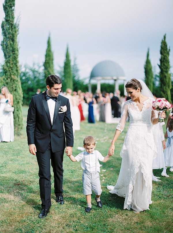 The bride and groom walk down the outdoor ceremony aisle immediately after their ceremony, holding the hands of a young ring bearer between them. The groom wears a black tuxedo with a black bow tie and a white floral boutonniere, while the bride wears a long-sleeved lace wedding gown with a V-neckline, a tiara, and a veil, and carries a bouquet of pink roses. The young boy is dressed in light gray shorts with suspenders and a white shirt. In the background, a domed circular gazebo or pavilion is visible flanked by tall cypress trees, with a large group of guests gathered behind them, including bridesmaids in blue dresses. The shot is a medium portrait taken at ground level, capturing the joyful recessional moment in a classic, formal style.