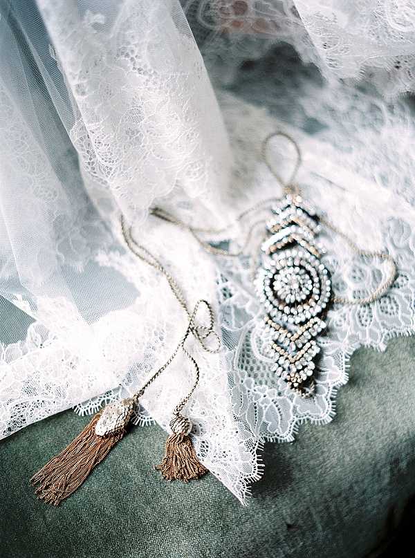 A close-up detail flat lay of bridal accessories arranged on a sage green velvet surface. A white lace-edged veil with scalloped trim is draped across the scene, alongside two gold chain tassel necklaces or pendants and an ornate beaded hair piece or brooch featuring crystal and pearl embellishments in a geometric, medallion-style design. The styling has a bohemian, vintage-inspired aesthetic with the combination of lace, gold metalwork tassels, and intricate beadwork.