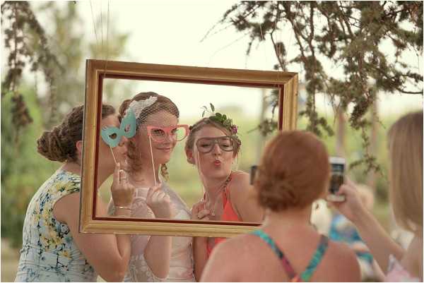 A group of four women pose at an outdoor photo booth station during what appears to be a wedding reception. The bride, identifiable by her white birdcage veil and white dress, holds a light blue masquerade mask on a stick while posing inside a gold ornate picture frame hung from a tree. A guest beside her wears a floral crown and holds oversized novelty glasses prop, making a puckered expression. A third woman in a coral dress stands nearby, while a fourth guest with red hair photographs the scene on a smartphone. The photo booth setup uses a large gold-painted frame as a fun prop, consistent with a boho or whimsical outdoor wedding style. Wide candid shot taken outdoors with soft natural light and greenery visible in the background.