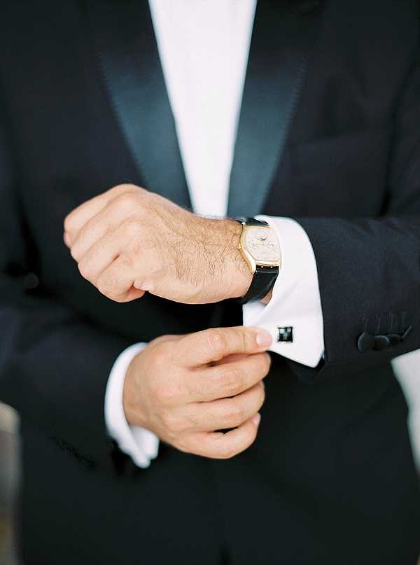 A close-up detail shot of a groom adjusting his cufflink while getting ready. He is wearing a black tuxedo with satin lapels, a white dress shirt with French cuffs, a dark cufflink, and a gold-cased dress watch with a black leather strap on his wrist. The composition is a tight torso-and-hands portrait with a clean, classic formal styling aesthetic.
