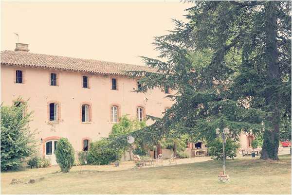 Wide exterior shot of a large pink-rendered French country house or mas, featuring terracotta roof tiles, shuttered windows with ochre surrounds, and a ground-floor covered terrace area. The image has a warm, faded vintage tone with soft muted hues. No people or wedding-specific decor are visible in the frame. Potential venue feature image.