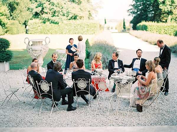 A cocktail hour or outdoor reception scene set in a formal French chateau garden, where approximately twelve guests are seated and standing around white wrought-iron tables and chairs arranged on a gravel terrace. The guests are dressed in formal evening attire — men in dark suits and black tuxedos with bow ties, women in cocktail dresses including a navy gown and a red dress. The tables are set with wine glasses and what appears to be a red patterned tablecloth on the central table. A large stone urn planter and manicured hedgerows frame the scene, with a chateau building and cypress trees visible in the background. The shot is a wide, slightly elevated angle capturing the full social gathering in its garden setting. Potential venue feature image.