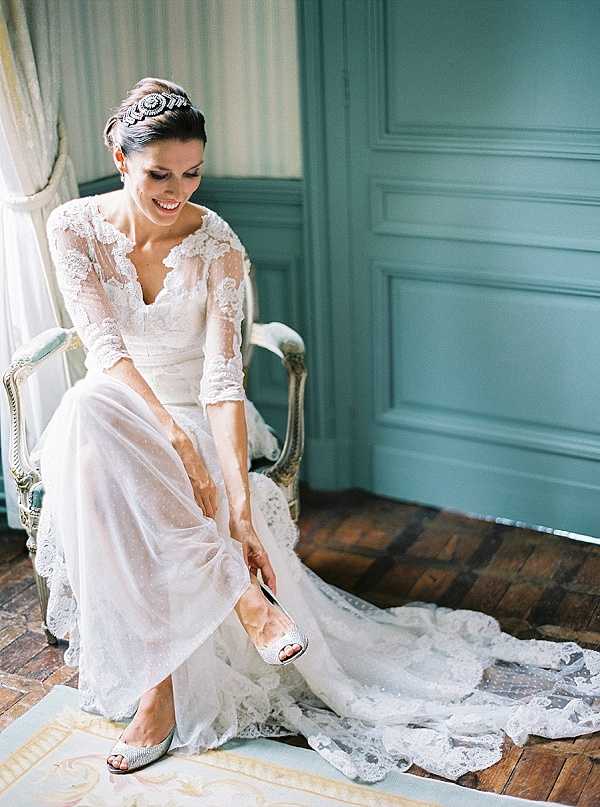 A getting-ready portrait of a bride seated in a white and gilt French-style armchair, putting on her shoes. She wears an ivory lace wedding gown with three-quarter-length sleeves, a deep V-neckline, and a long lace-trimmed train pooling on the herringbone parquet floor. Her hair is styled in an updo adorned with a crystal and rhinestone headpiece. Her shoes are embellished with silver or crystal detailing. The room features teal-painted wood-panelled walls and a curtained window, suggesting a classic French chateau interior. The shot is a medium portrait with a film photography aesthetic, warm and slightly soft in tone.