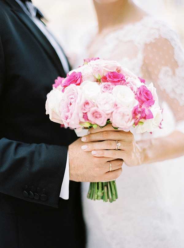 A close-up detail shot of a couple holding a bridal bouquet together, showing their hands and torsos only. The bouquet is a tightly arranged round composition of soft blush pink, hot pink, and white roses with green wrapped stems. The bride wears a lace long-sleeve gown with a scalloped lace overlay, and both wedding bands are visible — the bride's ring features a diamond solitaire on a gold band, and the groom wears a plain gold band. The groom is dressed in a dark navy or black suit jacket. The overall color palette centers on pink and white florals against classic black and white attire.