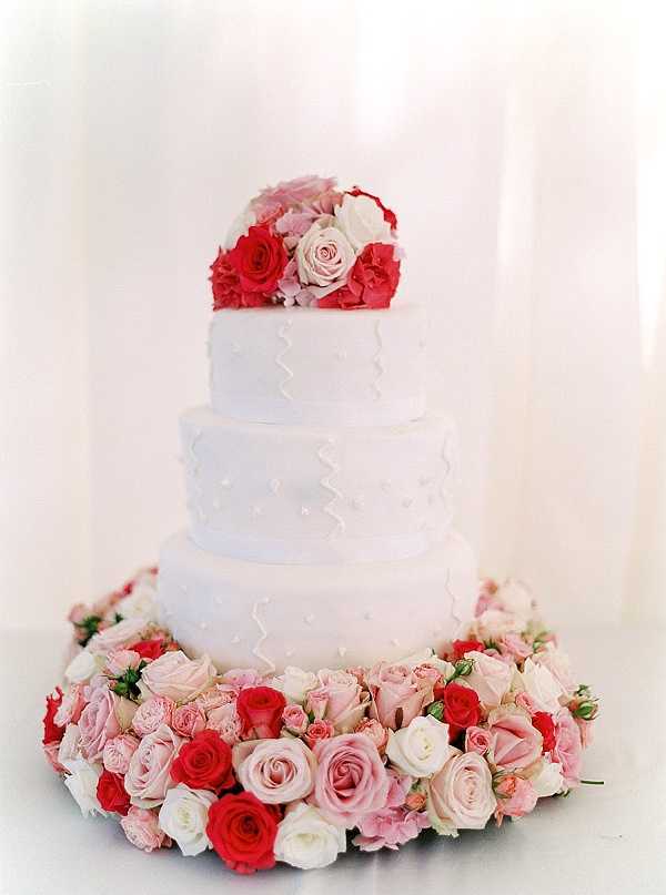 Close-up detail shot of a three-tiered white wedding cake decorated with a subtle zigzag piped pattern and small dot embellishments on each tier. The cake is surrounded at the base by a lush floral collar of blush pink, hot pink, red, and ivory roses, and topped with a matching cluster of the same blooms. The background is a soft, bright white, likely a draped fabric or wall. The overall decor palette is white and pink-to-red, with a classic, polished cake design style.