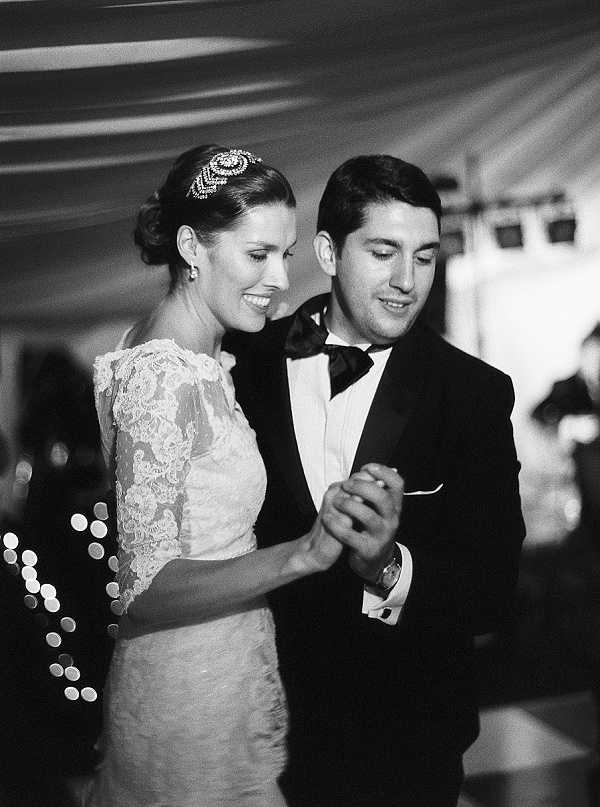 A black-and-white portrait-style shot of a couple sharing their first dance at an indoor reception, likely held under a draped tent or marquee. The bride wears a lace gown with elbow-length sleeves and an ornate beaded headpiece, with her hair in an updo; the groom is dressed in a classic black tuxedo with a bow tie and white pocket square. Their hands are joined as they dance closely together, both looking downward with relaxed expressions. Bokeh from what appear to be string lights or candles is visible in the background left, and stage lighting equipment is faintly visible in the upper right; the image has soft contrast with a fine-grain film quality.