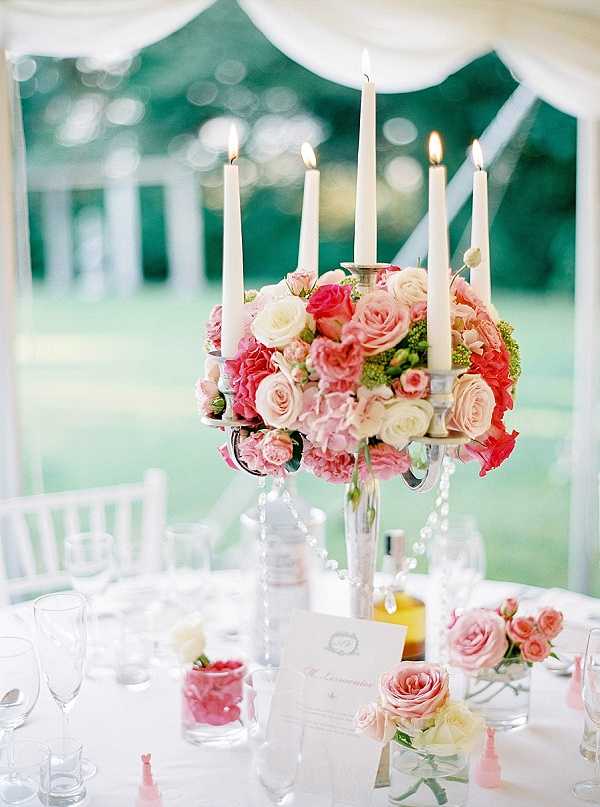 Close-up detail shot of a wedding reception centerpiece inside a white draped tent, with a lush floral arrangement of coral, hot pink, blush, and cream roses, pink hydrangeas, and small green buds mounted on a silver candelabra holding five lit ivory taper candles. Crystal bead garlands drape from the candelabra arms, and the white linen-covered table is set with stemmed glassware, a printed place card or menu, a small pink votive holder with rose petals, and a low secondary arrangement of blush and coral garden roses in a glass vessel. The overall decor palette is white, silver, blush, and coral with a classic, romantic style. The background reveals white chiavari chairs and a manicured lawn visible through the open tent sides.