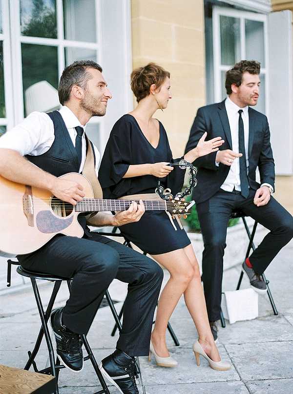 A three-piece live music band performs outdoors at what appears to be a wedding cocktail hour or reception, positioned in front of a classic French stone building. The guitarist on the left wears dark trousers, a black waistcoat, white shirt, and black tie; the female vocalist in the center wears a short black dress with nude heels and holds a tambourine; the male vocalist on the right wears a dark suit with a black tie. All three musicians are seated on black folding stools and appear mid-performance. The shot is a medium close-up portrait taken at a slight angle, with a film photography aesthetic visible in the warm, slightly faded tones.