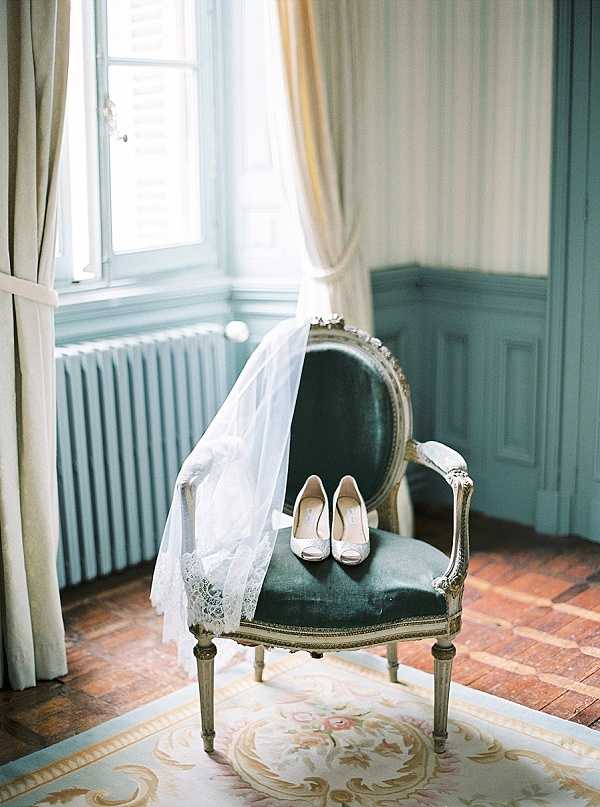 A bridal detail flat-lay-style shot taken indoors during a getting-ready moment, featuring a white lace-edged veil draped over the back and seat of an ornate Louis XVI-style armchair with a gilt wood frame and teal velvet upholstery. A pair of ivory peep-toe heels with a small bow detail are placed on the chair seat. The room features pale blue-grey wood-panelled walls, cream floor-length curtains, a white cast-iron radiator, terracotta tile flooring, and a floral Aubusson-style rug beneath the chair, all consistent with a classic French chateau interior. The composition is a medium wide shot emphasizing the chair as the central styling prop within the period room setting.