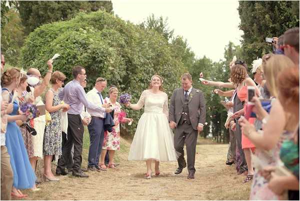 A newlywed couple walks through a confetti exit line outdoors on a dry dirt path, with approximately 25-30 guests lined up on either side throwing confetti. The bride wears a tea-length ivory dress with lace three-quarter sleeves and pink heeled shoes, while the groom wears a charcoal grey suit with a buttonhole. The bride carries or has passed off a bouquet featuring purple and blue flowers. Guests are dressed in summery attire including floral prints, bright colors, and occasion wear with fascinators. The scene has a relaxed, vintage-inspired feel with natural afternoon light. Wide shot capturing the full confetti exit moment.