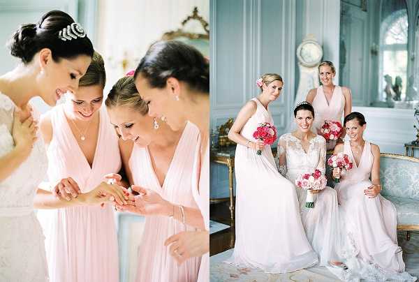 A two-image composite showing a bride with her bridal party during getting-ready and portrait moments inside what appears to be a French chateau with pale blue-grey paneled walls, ornate gold-framed mirrors, and classic period furniture. In the left image, the bride — wearing a white lace gown with an art deco crystal hair piece and an updo — huddles with three bridesmaids in blush pink deep V-neck draped gowns, all looking down at a piece of jewelry being examined; this is a close-up portrait shot. In the right image, the same bride sits on a pale upholstered antique sofa alongside three bridesmaids in the same blush pink gowns, all holding bouquets of hot pink and coral flowers including what appear to be roses and ranunculus; this is a medium group portrait shot. The overall styling palette combines ivory lace, blush pink, and vivid pink florals against the soft grey-blue classical interior.