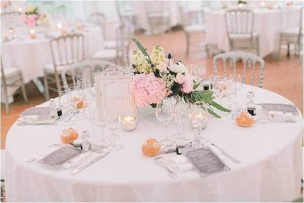 A close-up shot of a round reception table set for a wedding dinner in a bright, airy indoor venue. The table is covered with a white linen and set with wine glasses, gray folded napkins, and small clementines as decorative place setting accents. The centerpiece features pink hydrangeas, white ranunculus, yellow stock flowers, and green foliage in a low arrangement, framed by an ornate silver photo frame table number card. Small votive candles in glass holders are scattered across the table. The chairs are silver Napoleon-style. Additional similarly styled round tables are visible in the soft-focus background, indicating a spacious reception room with warm terracotta flooring. The overall decor palette is white, blush pink, and silver with a classic French style.