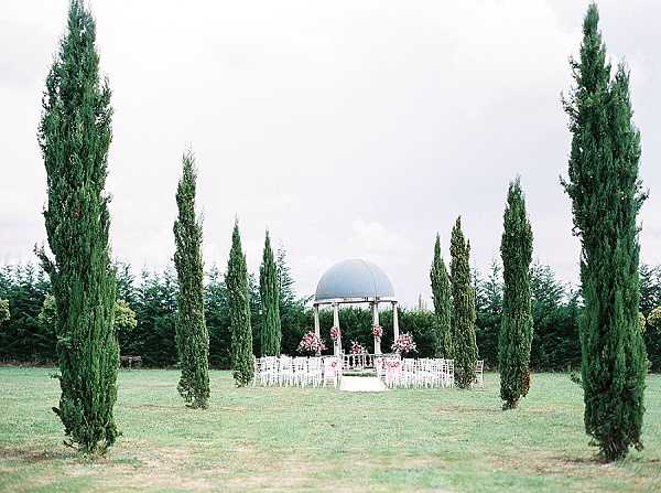 A wide shot of an outdoor ceremony setup centered around a white gazebo with a grey domed roof, positioned on a large lawn and framed symmetrically by tall Italian cypress trees. White chiavari chairs are arranged in rows on either side of a central aisle leading to the gazebo, which is decorated with pink and red floral arrangements at its columns. The overall styling is classic and formal, with the cypress tree arrangement giving a Mediterranean feel. No people are present in the image. Potential venue feature image.
