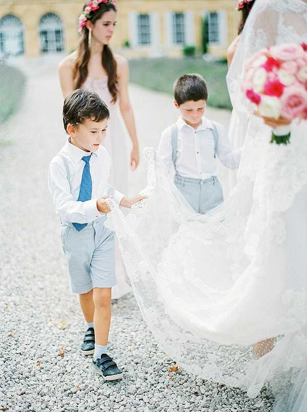 Two young page boys carry the lace train of a bride's wedding dress along a gravel path outdoors, with a yellow chateau facade visible in the background. Both boys are dressed in light blue shorts, white shirts, and suspenders, with one wearing a blue tie; they wear sneakers and white socks. A bridesmaid in a pale pink strapless dress with a pink floral flower crown stands behind them, looking downward. The bride, partially cropped on the right, holds a bouquet of pink and hot pink roses, and her lace-overlay gown features a long train. The shot is a candid medium portrait taken at ground level on a gravel path, capturing the children's focused expressions as they handle the train.