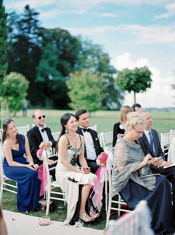 Guests are seated during an outdoor wedding ceremony, arranged in rows of white Chiavari chairs set on a lawn with open parkland in the background. Approximately six to eight guests are visible, including women in a cobalt blue one-shoulder gown and a black-and-white patterned dress, men in black tuxedos with bow ties, and an older woman in a silver-grey layered ensemble. The aisle chairs are decorated with hot pink carnation or peony pompoms tied with pink ribbon and pearl garland accents. The overall decor palette combines white, hot pink, and pearl tones, giving a classic formal style. Medium wide shot taken from a low angle looking along the seated rows toward the front of the ceremony.