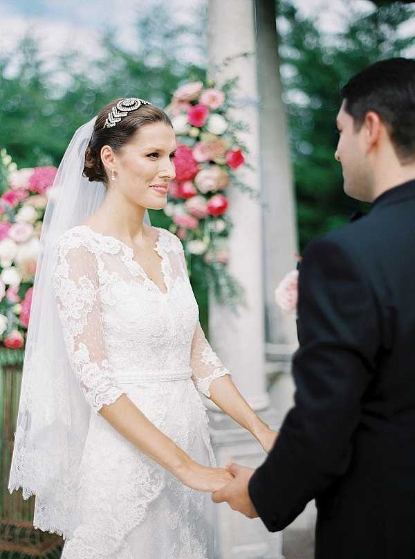 The bride and groom hold hands during an outdoor ceremony, captured in a close portrait shot from slightly above. The bride wears a white lace gown with three-quarter sleeves and a V-neckline, paired with a long veil and an ornate crystal headpiece worn across the crown of her upswept hair. The groom is dressed in a black suit and is shown from behind and to the side. Behind them, large floral arrangements in hot pink, blush, and white — appearing to include garden roses, peonies, and dahlias — are positioned against stone columns, suggesting a classic or formal outdoor ceremony setting. The overall styling is classic and formal with a rich pink and white floral palette.